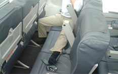 Photo of a man sitting by the window of an airplane, on a three-seat row, with his left leg stretched out on the two seats to his left.