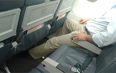 Photo of a man sitting by the window of an airplane, on a three-seat row, with his left leg stretched out under the front seat.