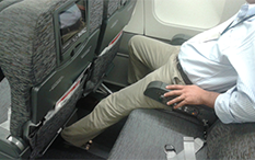Zdjęcie mężczyzny siedzącego przy oknie samolotu, w dwumiejscowym rzędzie, z lewą nogą wyciągniętą pod przednim siedzeniem.