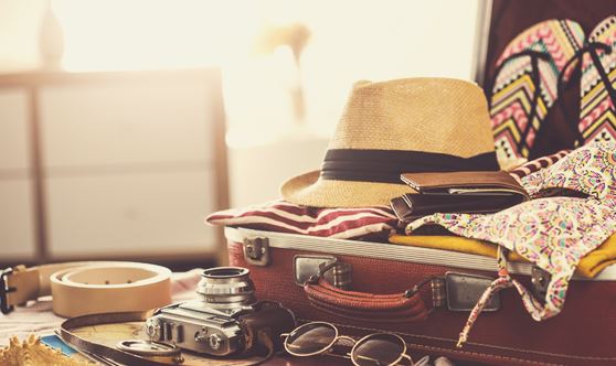 Composition with various objects related to the theme of travel, arranged next to and inside an open red travel bag. The objects include, among others: a straw hat, a colorful bikini top and a notebook.