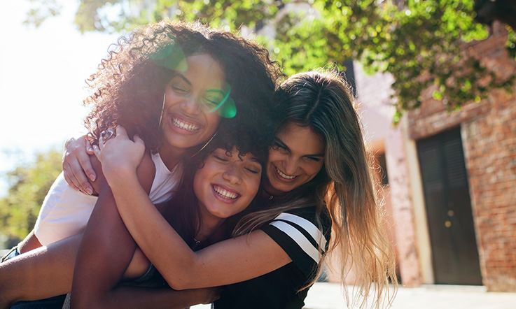 Image of 3 young women on the street. They are hugging and smiling. The young woman on the left is dark-skinned, has black curly hair, and is wearing a white tank top. In the middle there is another dark-skinned girl, but with straight hair. The girl on the right has straight blond hair and is wearing a black t-shirt. Behind them, slightly to the right, there is a rustic house. 