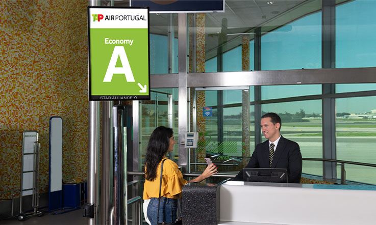 Widok punktu obsługi w strefie wejścia na pokład. Mężczyzna w ciemnym garniturze obsługuje panią w żółtej bluzce i dżinsach. Kobieta trzyma telefon komórkowy prawą ręką i pokazuje jakieś informacje. Obok biurka znajduje się szyld z logo TAP Air Portugalia, a pod nim napis „Economy” oraz duża litera „A” wyróżniona na zielonym tle.