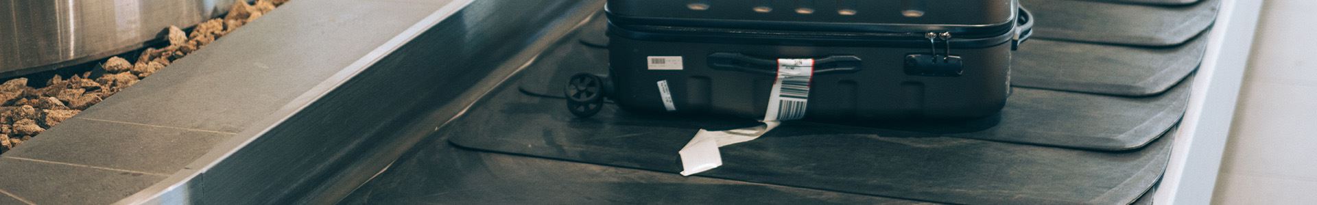 Imagem de uma mala de mão preta, rígida, com alças superiores e laterais, e com uma etiqueta de bagagem branca na alça lateral, deitada em uma esteira de bagagem.