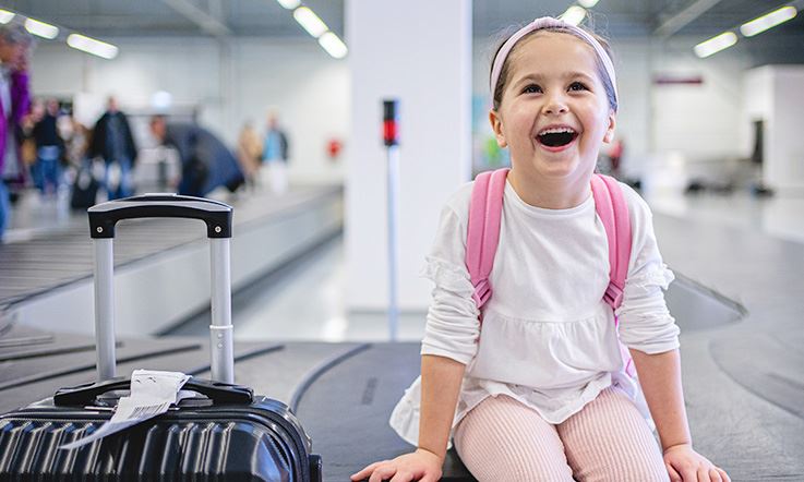 Улыбающийся ребенок с чемоданом рядом с ней сидит у подножия ленты для сбора багажа в аэропорту. Комната освещена, а на заднем плане несколько человек забирают свои вещи.