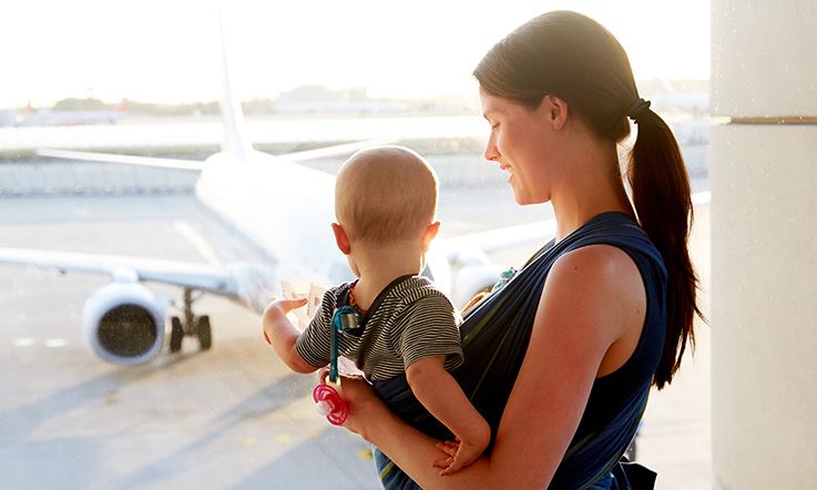Женщина, обращенная лицом в сторону, держит ребенка на коленях, спиной к изображению. Они в аэропорту и смотрят из окна на самолет на взлетно-посадочной полосе, при дневном освещении.