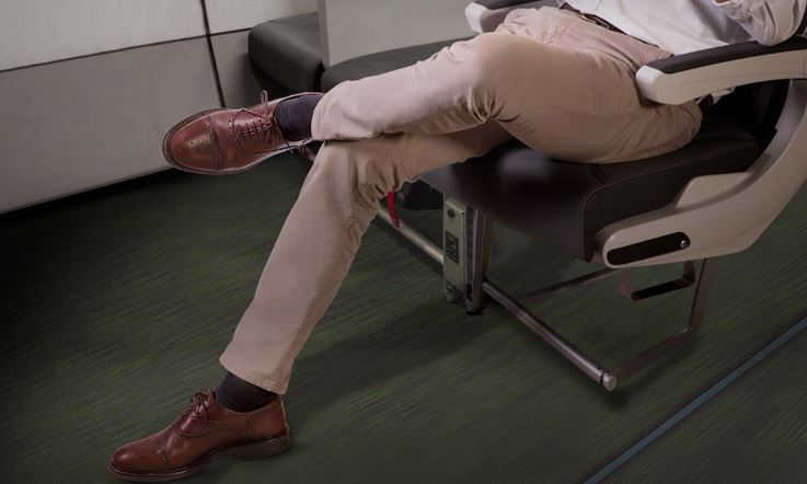 Primer plano de un hombre, de la cintura para abajo, sentado en un asiento de emergencia. Tiene las piernas cruzadas y viste pantalones de traje color beis y zapatos marrones.