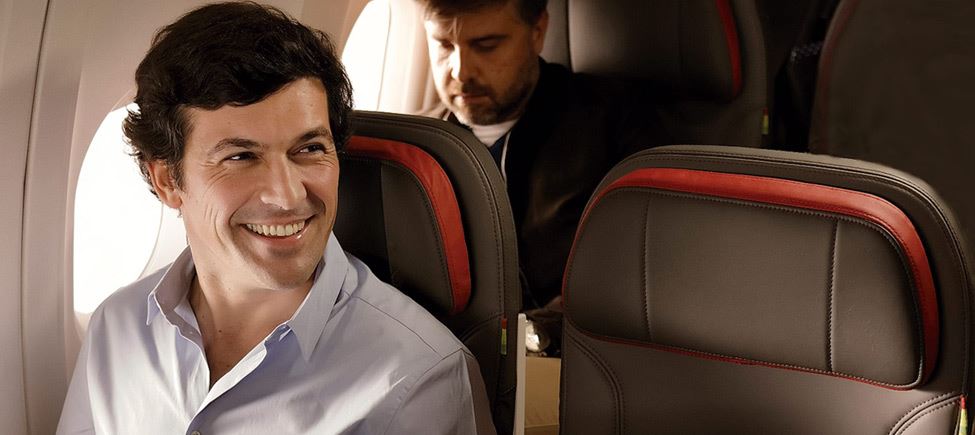 Gros plan d'un homme assis près de la fenêtre d'un avion. Il est brun et porte une chemise bleu clair. Il sourit et regarde sur son côté gauche.