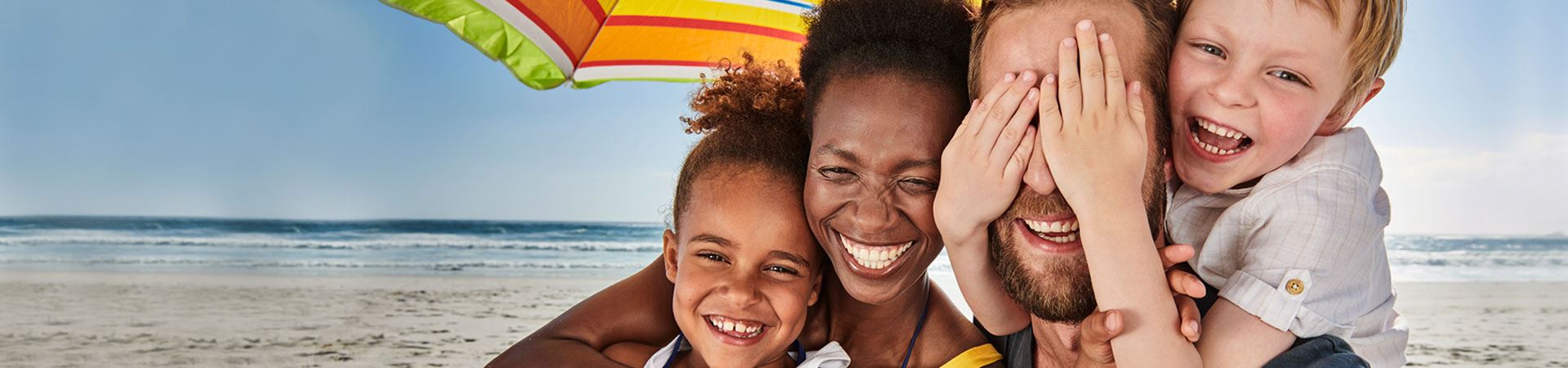 Das Bild zeigt eine Familie am Strand, bestehend aus einem Mann, einer Frau, einem Jungen und einem Mädchen. Sie sind alle zusammen unter einem Sonnenschirm, mit lächelnden und glücklichen Gesichtern. Das Mädchen wird von der Frau umarmt, während der Junge mit seinen Händen die Augen des Mannes zuhält. Im Hintergrund ist das Meer zu sehen, am Ende eines Sandstreifens.