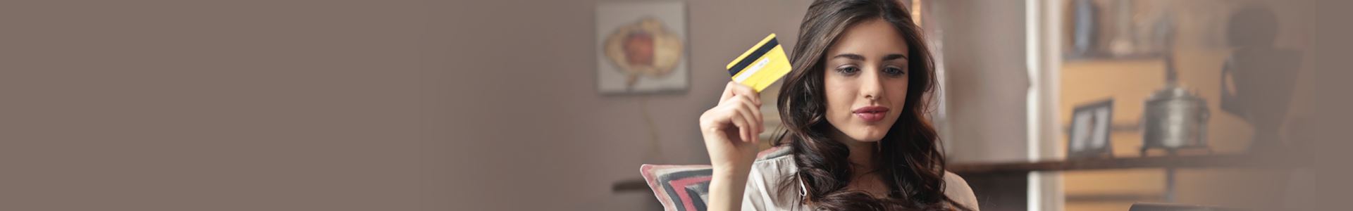 A brunette woman is sat leaning against a cushion and holding a yellow credit card in her right hand. She makes a small smile.