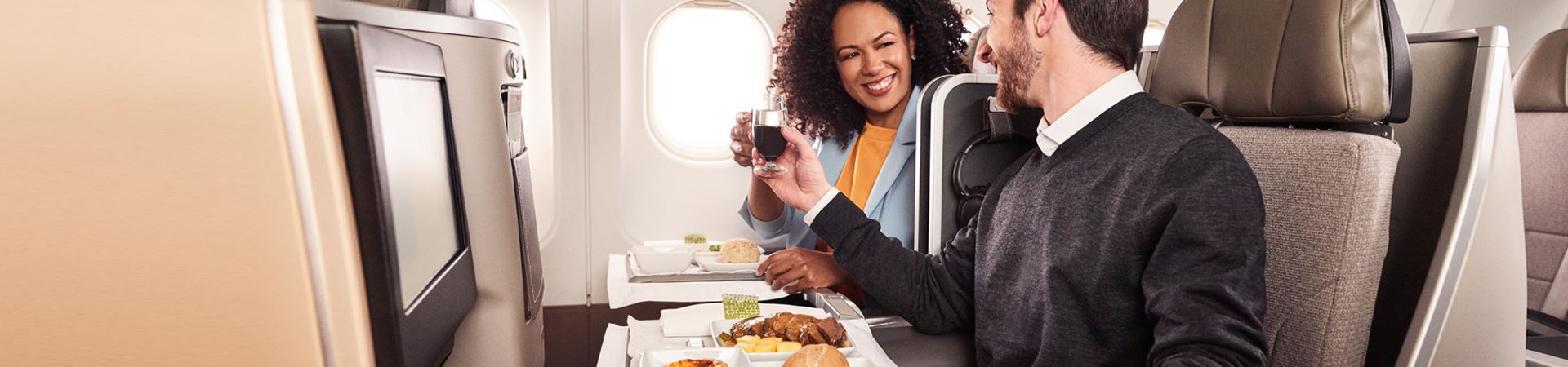 A smiling man and woman sitting side by side aboard a TAP plane. Both hold a glass in their hand and make a toast. In front of each, there is a tray with a meat meal, a bread and a "pastel de nata".