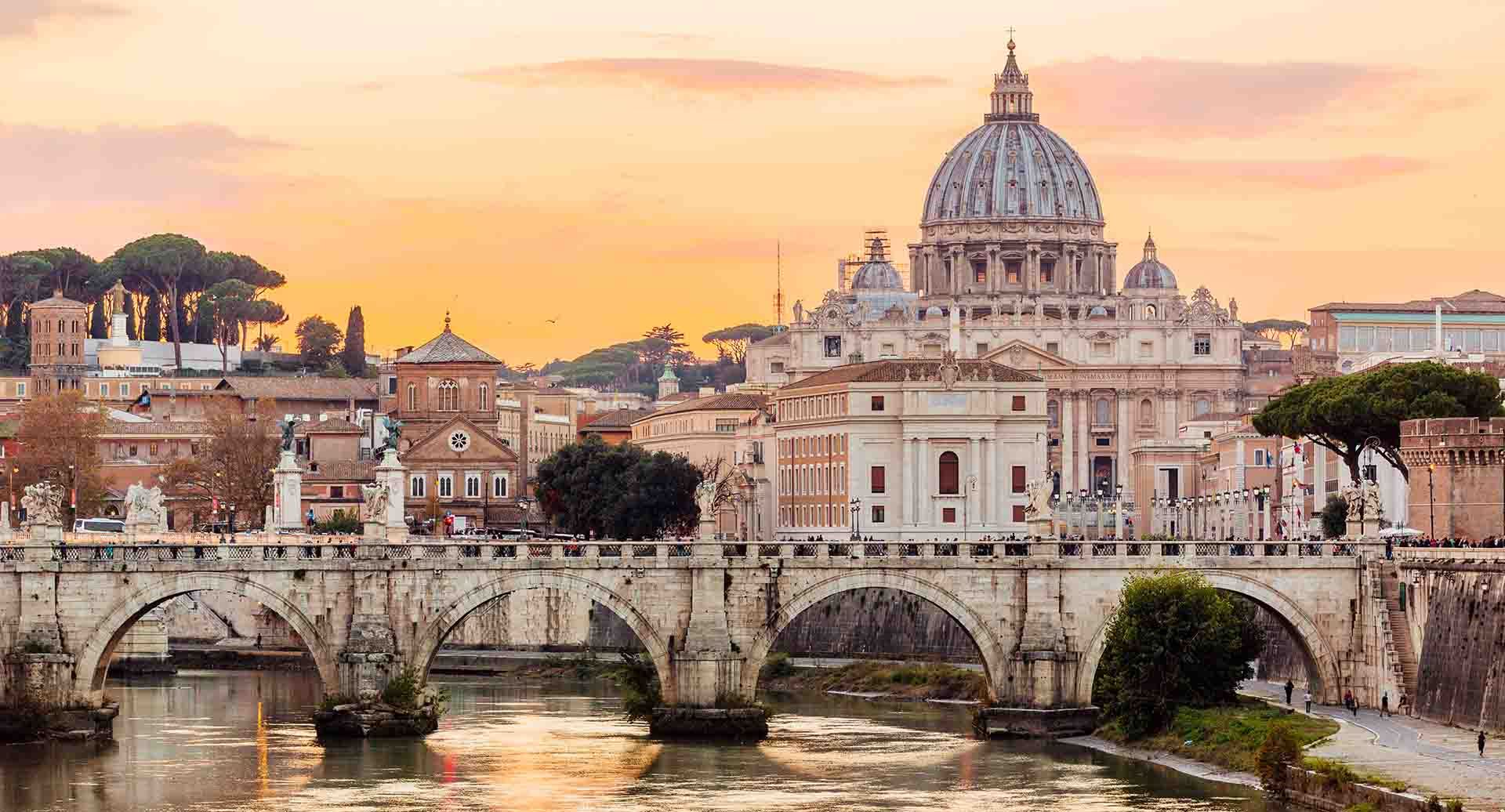 Picture showing a river in Rome and, in the background, a white stone bridge with arches through which the river is flowing; behind it, there are several buildings, a sunset, and an orange sky.