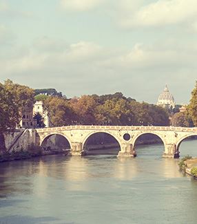 Photo of the Tiber River in Rome, Italy. One can see the river flanked by walls and trees with branches hanging over those walls. In the middle of the river there is a light-colored bridge with four arches. In the background one can see a dome and a blue sky with some white clouds.