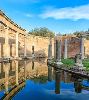 Photo of an area of Villa Adriana in Tivoli, Italy. The photo shows the ruins of an old building. One can see a ruined, incomplete, roofless brick wall. In the center, a water stream is flanked by several ancient columns, some of which are intact, while others are not as tall as expected. Behind the columns, on the right side, one can see a small lawn.