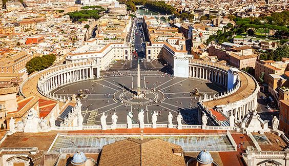 Panoramic photo taken from a high angle, focusing on Saint Peter's Square, Vatican, Italy. At the end of the street leading to the square there is a river with two bridges. Next to it, one can see the roof of several houses and some green areas with trees and gardens. The sky is blue, with a few white clouds.