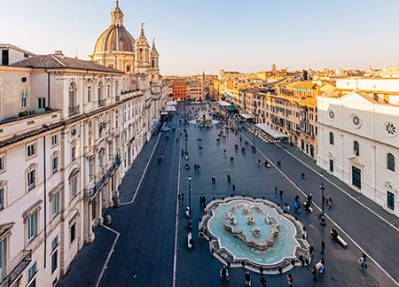 Aerial view of Piazza Navona in Rome, Italy. The square's ground is dark. One can see two fountains; the one in the background has a monument inside. The square's street is lined with light-color buildings and store awnings. At the end of the street, to the left, one can see the dome of the Church of Sant'Agnese in Agone.