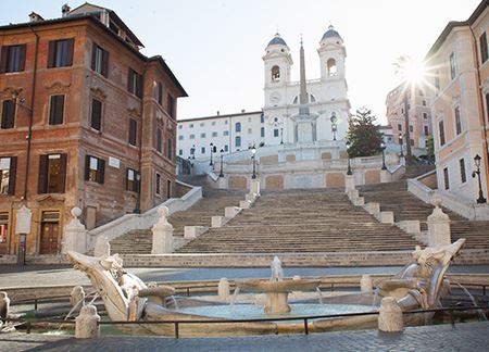 Photo of the Spanish Steps in Rome, Italy. In the foreground, there is a water-filled fountain in the center of a round piazza. Behind it, one can see a staircase flanked by two buildings and the church of Trinità dei Monti on top.
