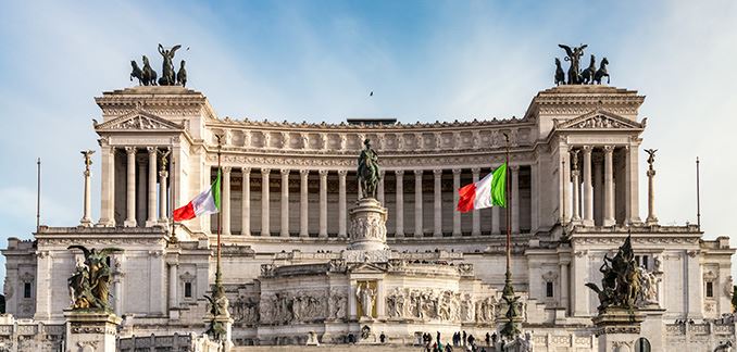 Front view of the Vittoriano monument in Rome, Italy. At the base of the monument there is a wide staircase, initially flanked by two pillars with two statues depicting three men and an angel, and at the end by two pillars bearing Italy's green, white and red flag. After the staircase, in the center, one can see a large statue of a man riding a horse. Behind the statue there is a corridor of pillars supporting the top of the building, which features, on both the right and the left sides, two statues representing four horses and an angel.