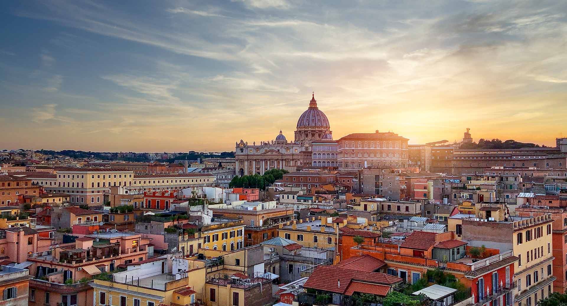 Imagem constituída por um plano aéreo da cidade de Roma. Em primeiro plano temos vários edifícios habitacionais e, em plano de fundo, vários monumentos da cidade de Roma com o pôr-do-sol por trás.
