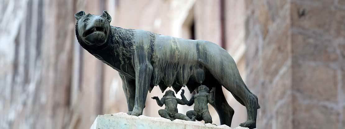 Low-angle picture of a bronze sculpture depicting a she-wolf standing on a white stone pulpit while suckling two naked children that are sitting below it. There is a beige stone Roman building In the background.