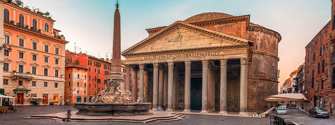 Side picture of a street showing the Rome Pantheon and a fountain in the background. The side streets in the picture consist of orange buildings and cobbled roads with cars.