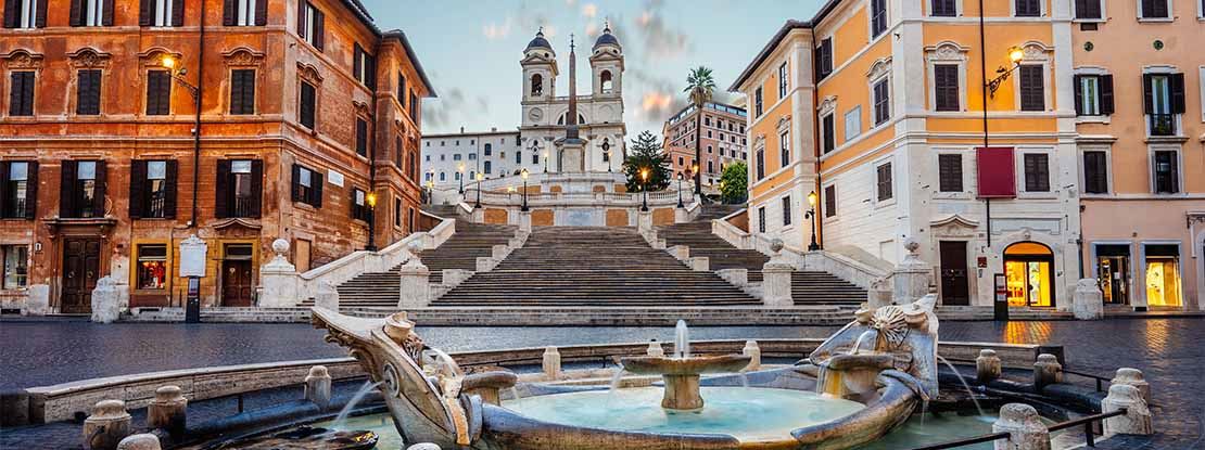 Picture of a street in Rome showing a fountain in the foreground and a white stone staircase flanked by two orange buildings in the background. At the bottom of the stairs there is a white stone building that resembles a church with two towers.