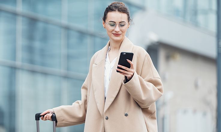 Fotografia de uma mulher usando óculos e um casaco quente de inverno, que sorri para o ecrã do smartphone que segura na mão esquerda, enquanto a mão direita está agarrada à pega de uma mala tipo trolley.