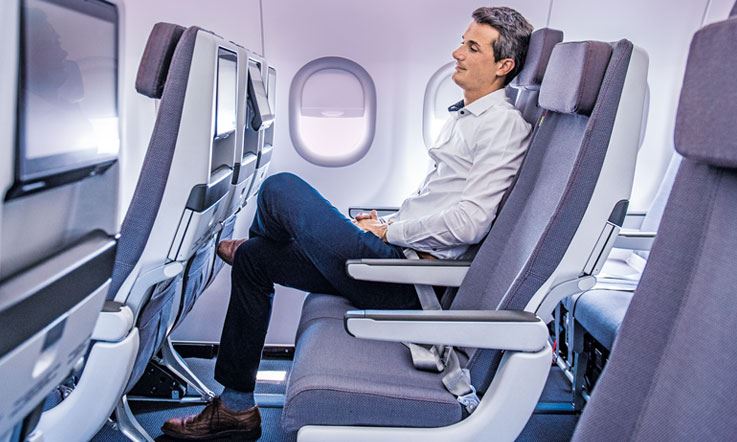 Фотография мужчины, одиноко сидящего на одном из 3 кресел в ряду самолета. Мужчина сидит в центре ряда кресел, положив левую ногу на правую. На нем синие брюки и белая рубашка. 