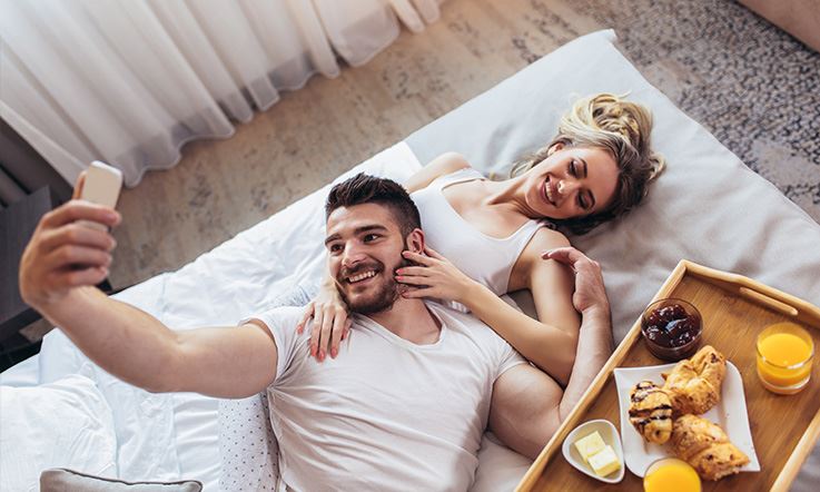 Zdjęcie mężczyzny i kobiety leżących na łóżku w pokoju hotelowym. Mężczyzna leżąc z głową na brzuchu kobiety trzyma telefon komórkowy tak, jakby robił im selfie. Obok pary znajduje się taca z rogalikami, sokiem pomarańczowym, masłem i dżemem. 