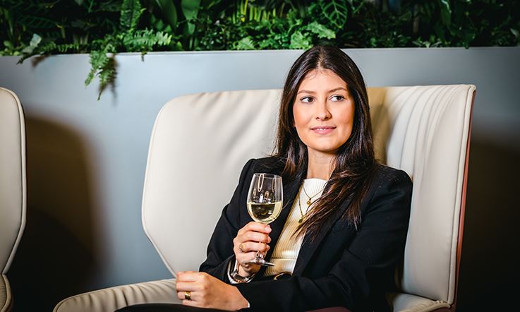 Фотография брюнетки, сидящей в шезлонге с бокалом вина в руке. На женщине черный блейзер поверх белой рубашки. 