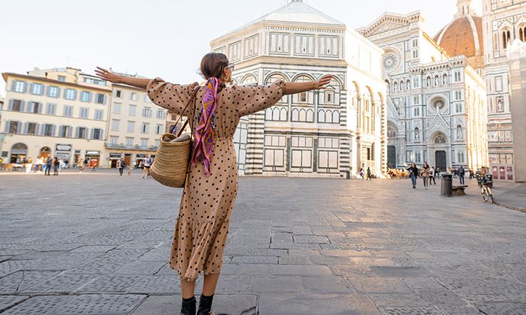 Zdjęcie kobiety z szeroko rozłożonymi ramionami na placu katedralnym we Florencji. Kobieta zwrócona jest twarzą do katedry i ma na sobie długą suknię w kwiaty. 