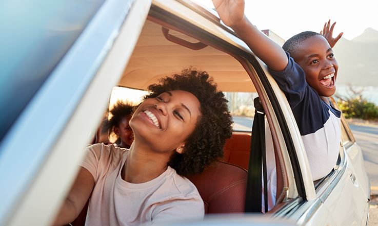 Zdjęcie kobiety i dwójki dzieci w białym samochodzie z otwartymi oknami. Kobieta uśmiecha się na siedzeniu kierowcy. Na tylnym siedzeniu jedno z dzieci wychyla się przez okno wyciągając ramiona i uśmiechając się. 