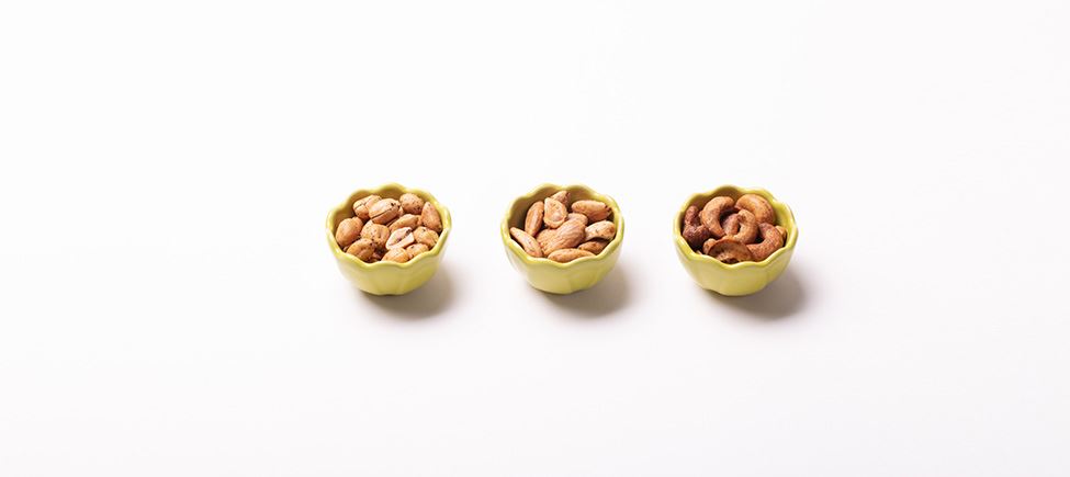 Photography of three green porcelain bowls on a white background. Each bowl contains a different nut - the first contains peanuts, the second almonds and the third cashews.