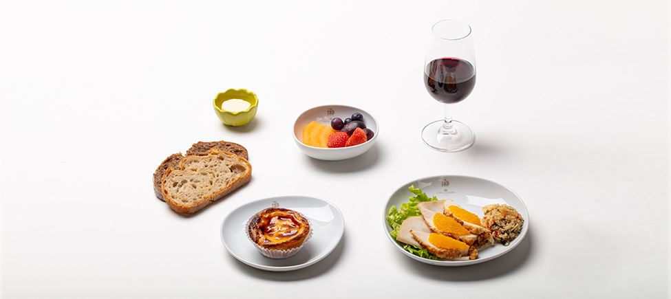 Komposition mit verschiedenen Elementen: zwei Scheiben Brot, eine Butterdose, eine Schüssel mit verschiedenen Früchten, ein Teller mit einem Pastel de Nata, ein hohes Glas mit Rotwein und ein Teller mit geschnittenem Lendenfilet, Salat, Orangen und Quinoa.