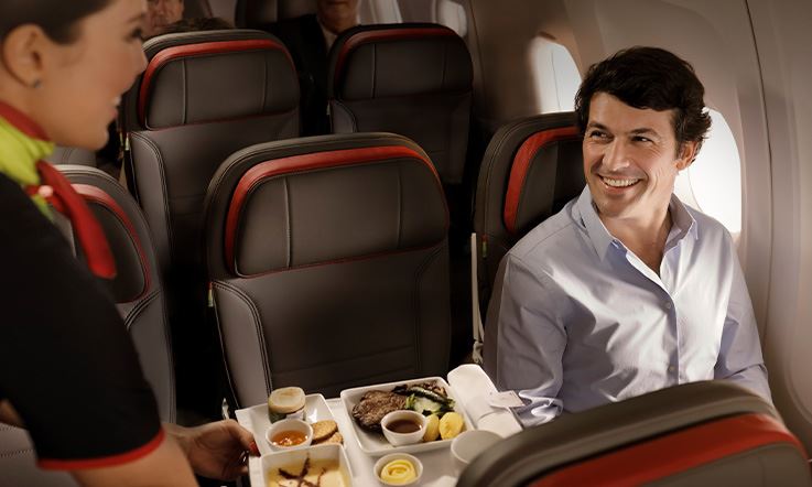 Foto de um homem sentado dentro de um avião perto da janela. O homem olha e sorri para uma aeromoça, que está do lado esquerdo da imagem, e segura uma bandeja com vários pratos de comida.