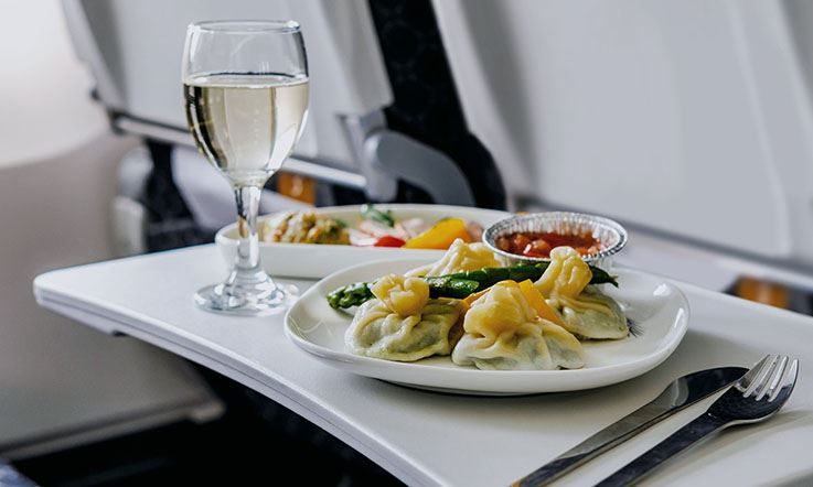 Fotografia de uma refeição - dois pratos de comida, um copo alto de vinho branco e alguns talheres - na mesa lateral de uma poltrona de avião.