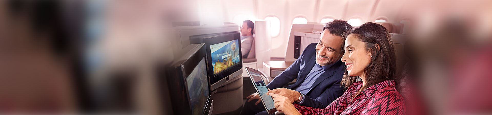 Close up of a smiling woman and a smiling man sitting on an airplane. The woman is holding a tablet, and the man is looking at it. They are both pointing to the tablet screen. There are two screens in front of them, built into the plane's seats.