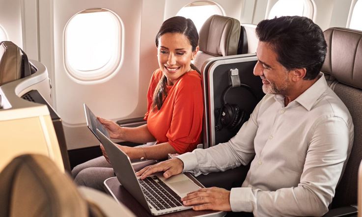 Some passengers who are a couple, are sitting in more spacious and comfortable chairs in Executive Class, in an environment with plenty of natural light, and without people around them. The male passenger is happy while typing on his computer, on the backup table of his chair. The female passenger is smiling, with a tablet in her hands, while looking at the passenger's computer.