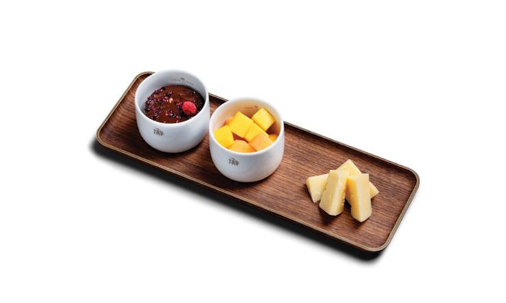 Fotografia drewnianej tacy, na której stoją dwie białe miski, a po prawej stronie znajdują się cztery kawałki sera. W misce po lewej stronie znajduje się deser z czekolady i malin, a w drugiej misce jest pokrojone w kostkę mango.