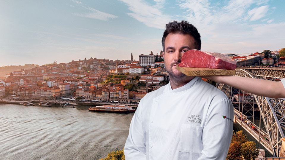 Medium shot of Chef Arnaldo Azevedo. He wears a white chef's coat with "The art of tasting Portugal" written on the upper left pocket, on his chest, and the TAP Air Portugal logo on his left arm. Behind the Chef, in the background, is a panoramic photograph of Porto with the Douro River and the city in the background and the D. Maria Pia Bridge on the right. Also to the right is an arm holding a piece of Maronesa meat in front of the Chef, partially covering the left side of his face. 