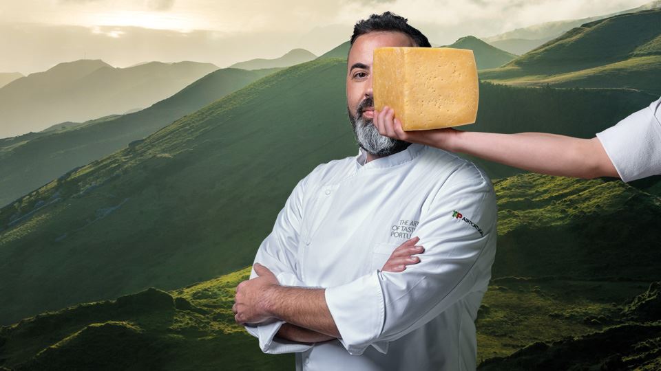 Medium long shot photo of Chef Cláudio Pontes dressed in a white chef jacket, with his arms crossed over his chest. To the right, a hand is holding a piece of São Jorge cheese in front of the Chef's face. In the background, a panoramic photo of São Miguel island's lush green hills.
