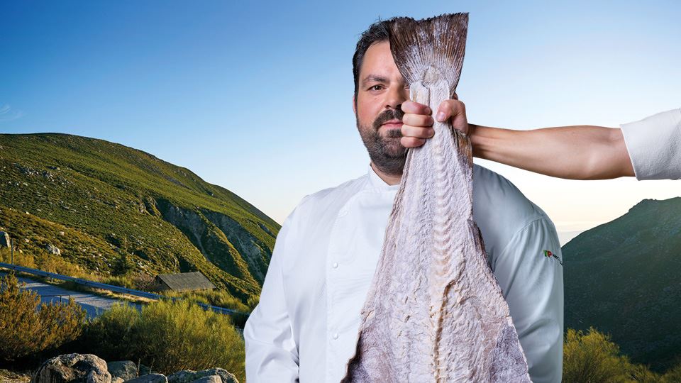 Medium shot of Chef Diogo Rocha, dressed in a white kitchen coat. On the right hand side, an arm appears holding a dried salted cod in front of the Chef, partially covering the left side of his face. In the background, there is a panoramic photograph of the hills full of green vegetation in central Portugal.
