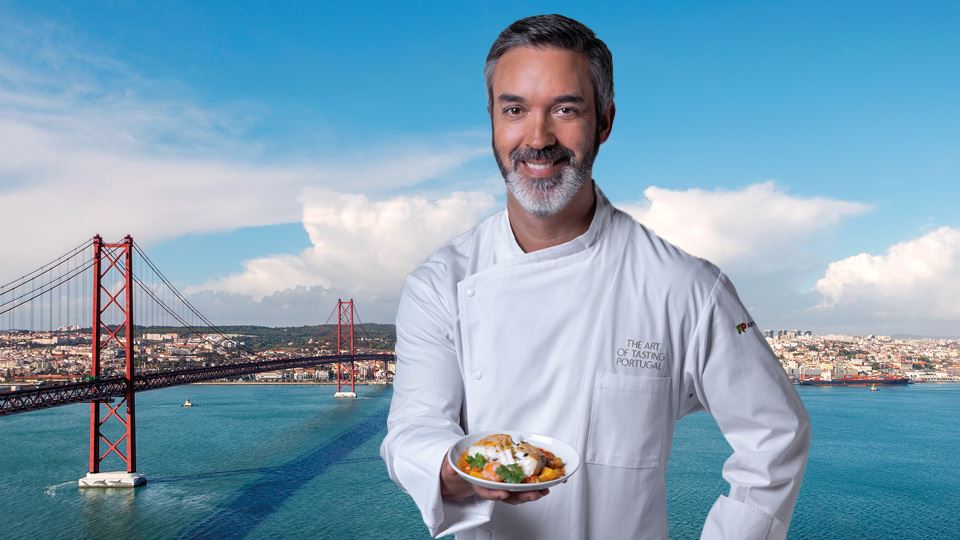 Medium shot of Chef Henrique Sá Pessoa smiling, dressed in a white chef's coat and holding a unique dish with grouper. In the background there is an aerial image of Lisbon and the Tagus river, with the 25th April bridge on the left side.