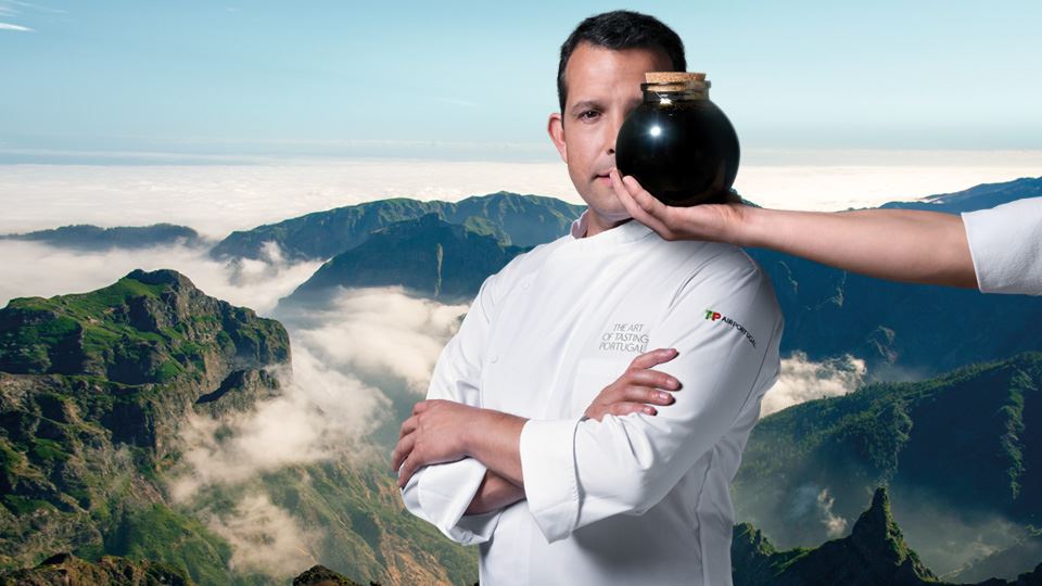 Foto panorámica de las exuberantes montañas de Madeira Island. A la derecha, superpuesto a la foto, el Chef Júlio Pereira sostiene una botella de Madeira Sugarcane Molasses con su mano izquierda. La botella cubre la parte inferior izquierda de su cara.