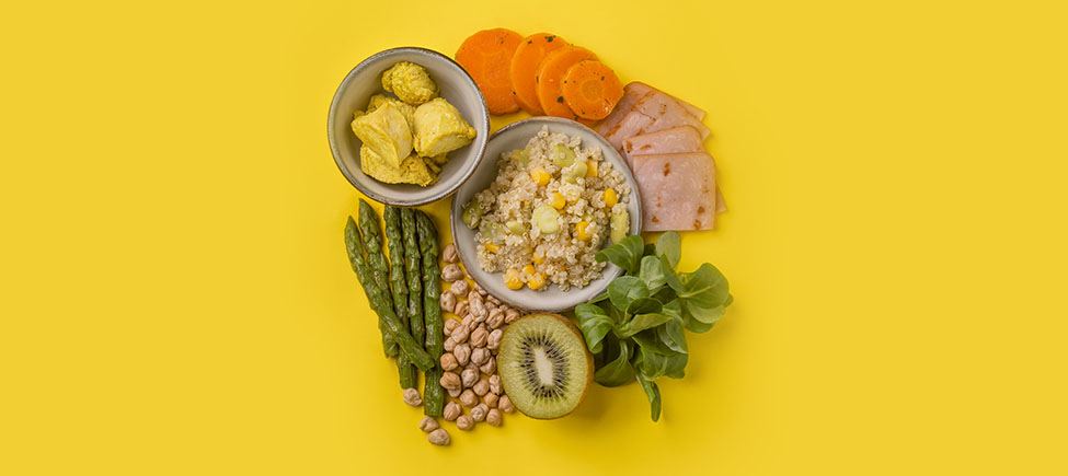 Фотография с различными продуктами, представляющими диету с низким содержанием натрия, на желтом фоне: спаржа, злаки, киви, овощи, морковь и другие продукты, подходящие для этого типа питания.
