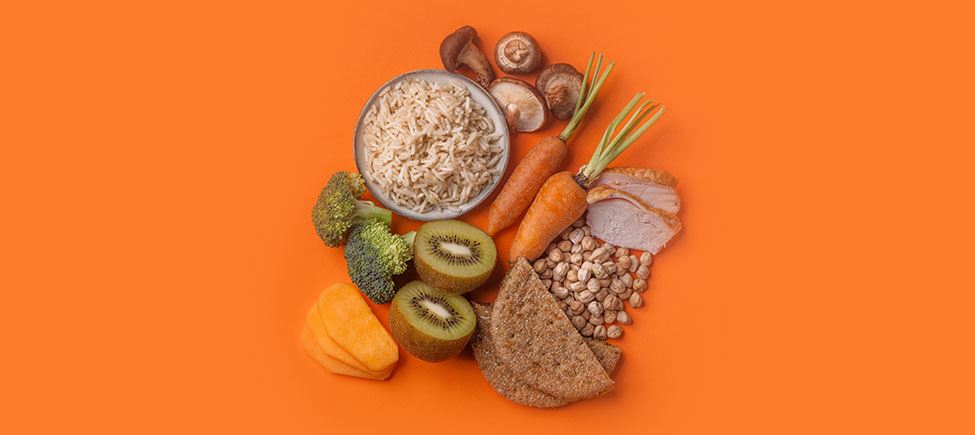 Фотография с различными продуктами, представляющими диету для диабетиков, на оранжевом фоне: грибы, рис, брокколи, морковь, киви, крупы, белое мясо и другие продукты, подходящие для этой диеты.
