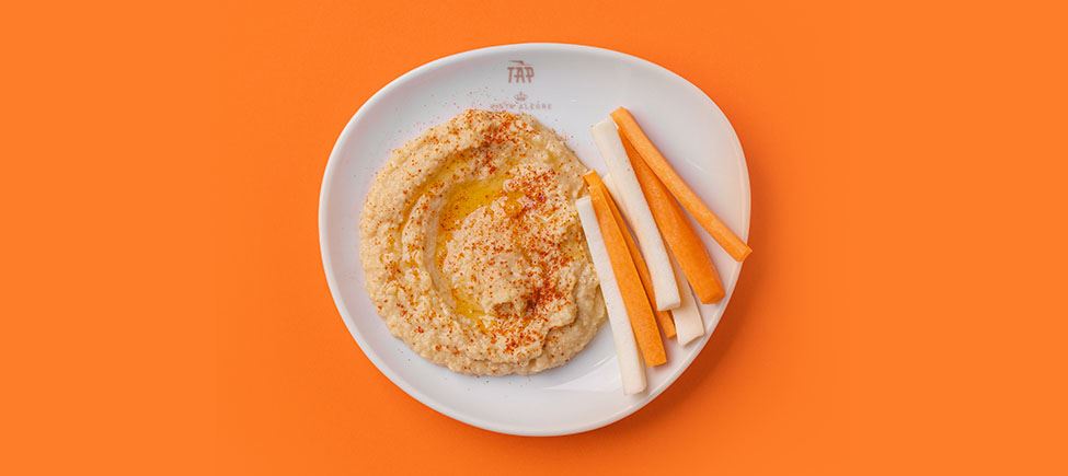 Une photo d'une assiette blanche avec du houmous et des bâtonnets de légumes sur fond orange.