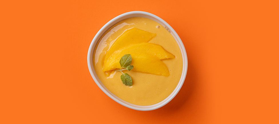 Photo of a white bowl with a mango mousse topped with a mint leaf, against an orange background. 