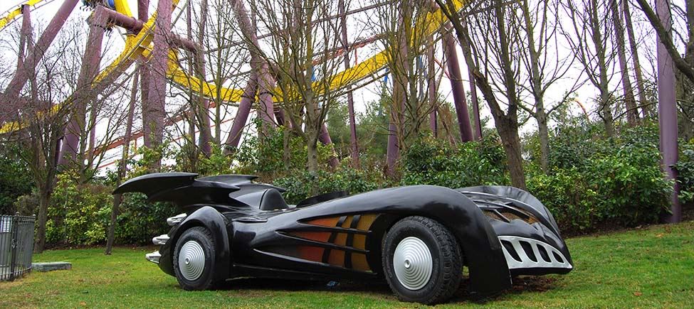 Фотография машины, похожей на машину Бэтмена, в натуральную величину. Он черный и с низкой посадкой, с неровными боками и двумя черными крыльями на спине. Стоит в саду.