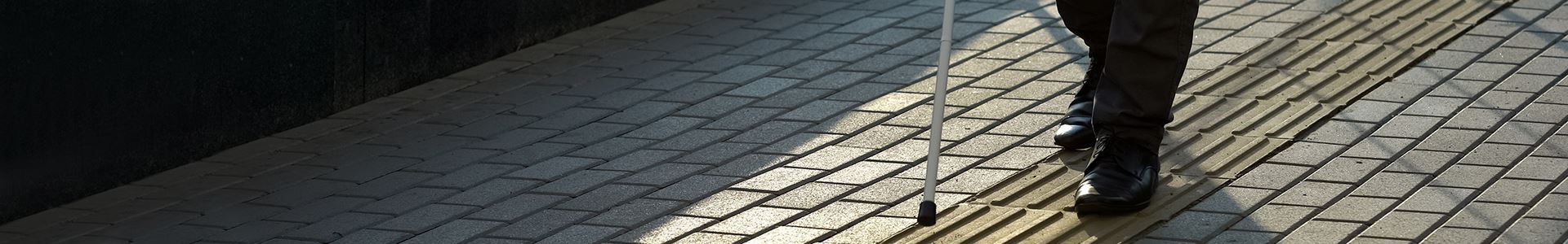 Abbildung eines Unterkörpers mit schwarzer Hose und schwarzen Schuhen und mit einem Blindenstock für Sehbehinderte auf einem grauen Bürgersteig mit einer ertastbaren Bodenleiste für Sehbehinderte.