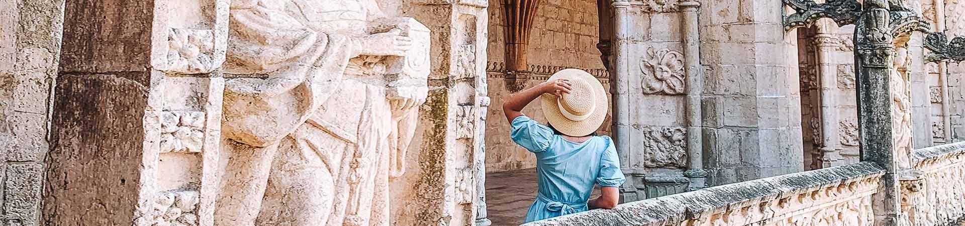Imagem composta por uma mulher vista de costas enquanto contempla um monumento de pedra branca. A mulher tem longos cabelos negros e está usando um vestido azul e segurando um chapéu de palha bege na mão esquerda.